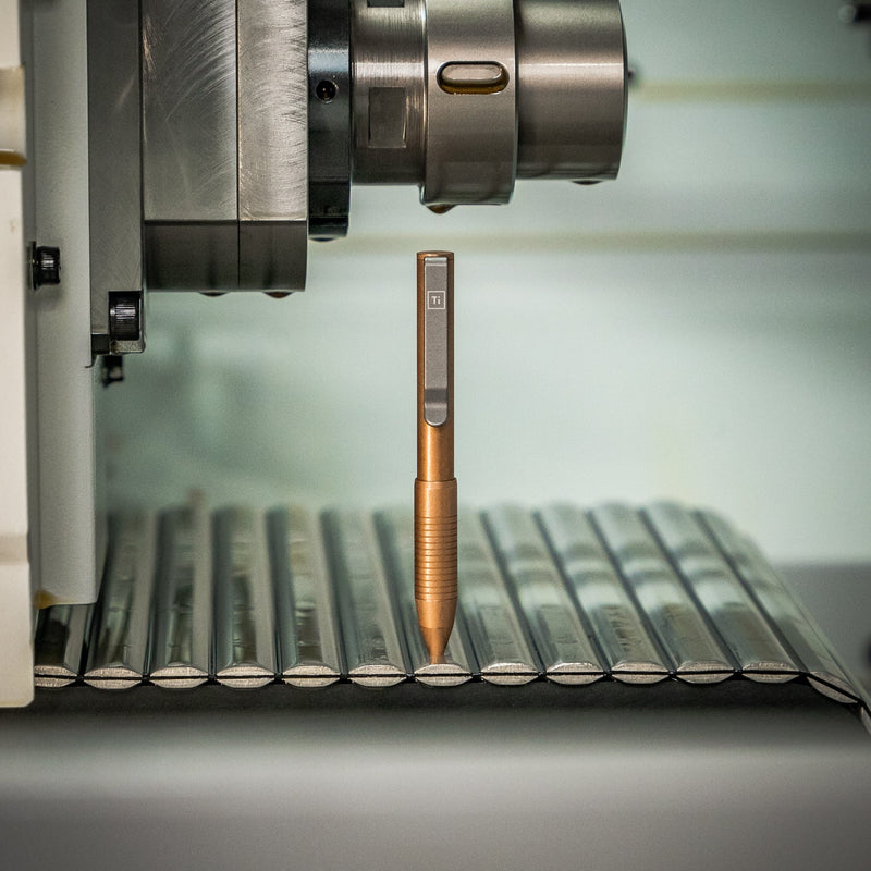 Bronze EDC pen made in USA standing vertically on metal surface under industrial machine