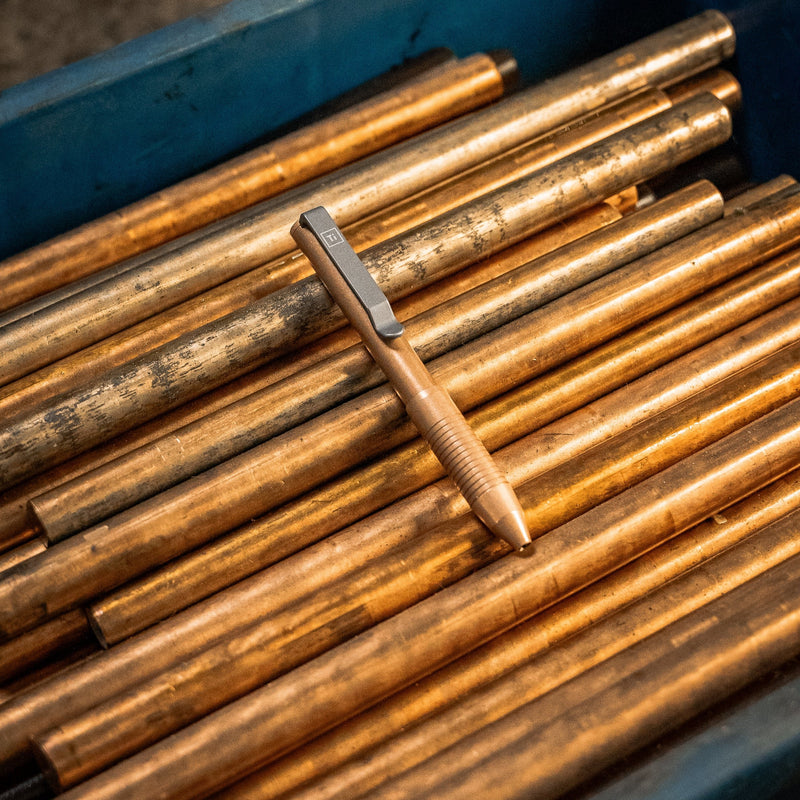 Bronze EDC pen made in USA resting on bronze metal rods in a blue container