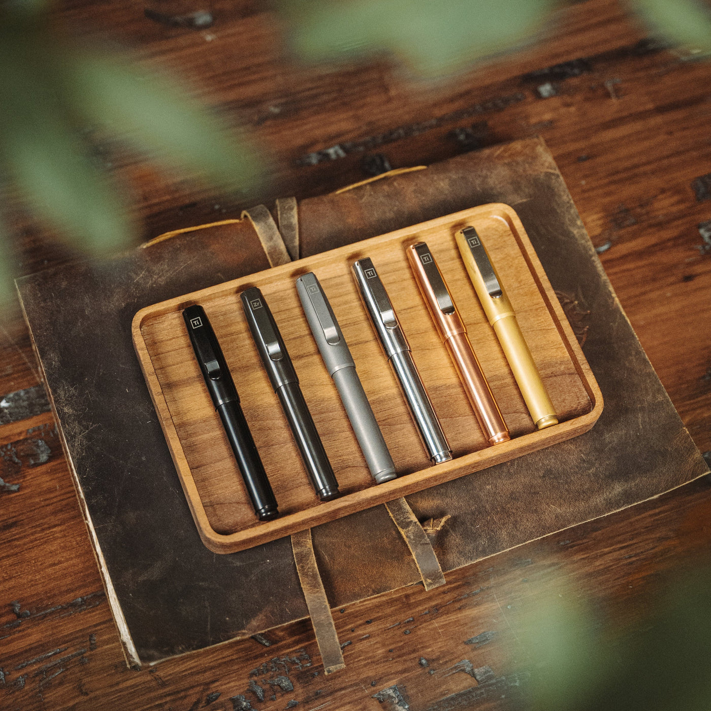 Big Idea Design LLC fountain pen for EDC enthusiasts in black, gunmetal, silver, copper, and gold finishes on wooden tray