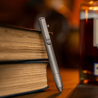Big Idea Design LLC EDC bolt action pen standing against old books on a wooden surface