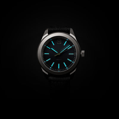 Big Idea Design LLC titanium field watch with luminous blue hands and hour markers on black dial and fabric strap