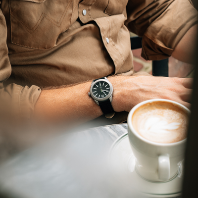 Big Idea Design LLC titanium field watch on wrist with black dial and leather strap by coffee cup