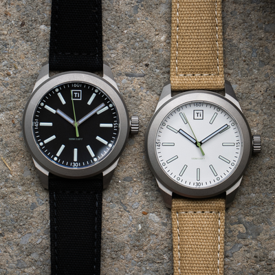 Big Idea Design LLC titanium field watch pair showing black dial with black strap and white dial with tan strap on concrete