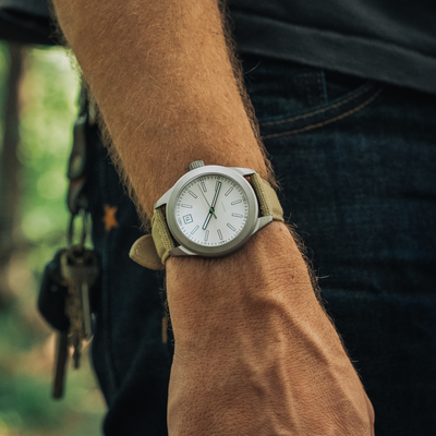 Big Idea Design LLC titanium field watch on a wrist with khaki canvas strap and white dial outdoors