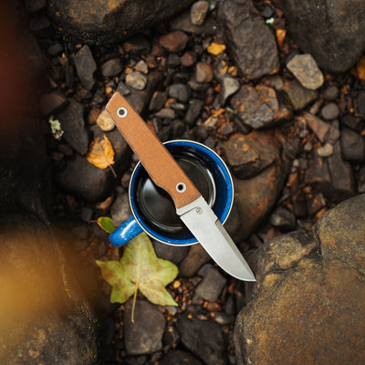 BIGiDESIGN-USA Fixed Blad knife with brown handle resting on blue mug outdoors on rocky ground