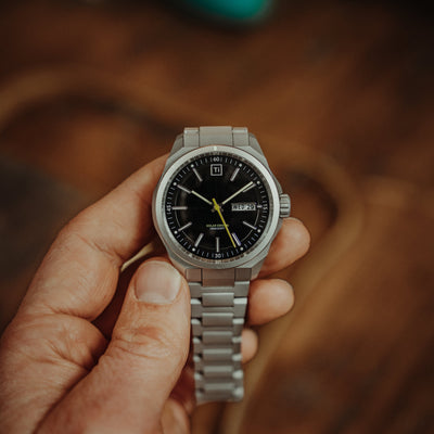 Hand holding a watch with a premium titanium bracelet and black dial showing time and date
