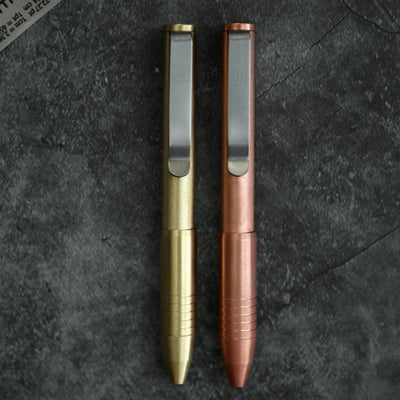 Two compact brass and copper metal pens with pocket clips by BIG IDEA DESIGN LLC on dark stone background