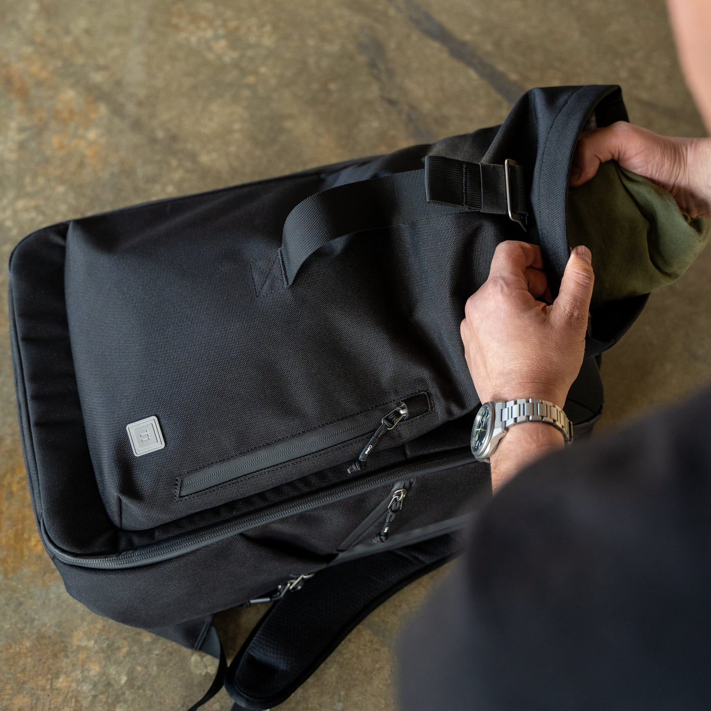 Big Idea Design LLC black everyday carry backpack with roll-top and front zipper pocket being packed by user