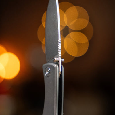 Close-up view of Big Idea Design LLC titanium pocket knife blade and handle with blurred warm lights background