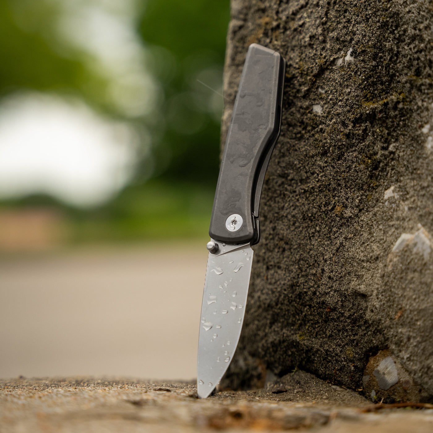 Titanium pocket knife with water droplets standing upright against a rough stone surface outdoors