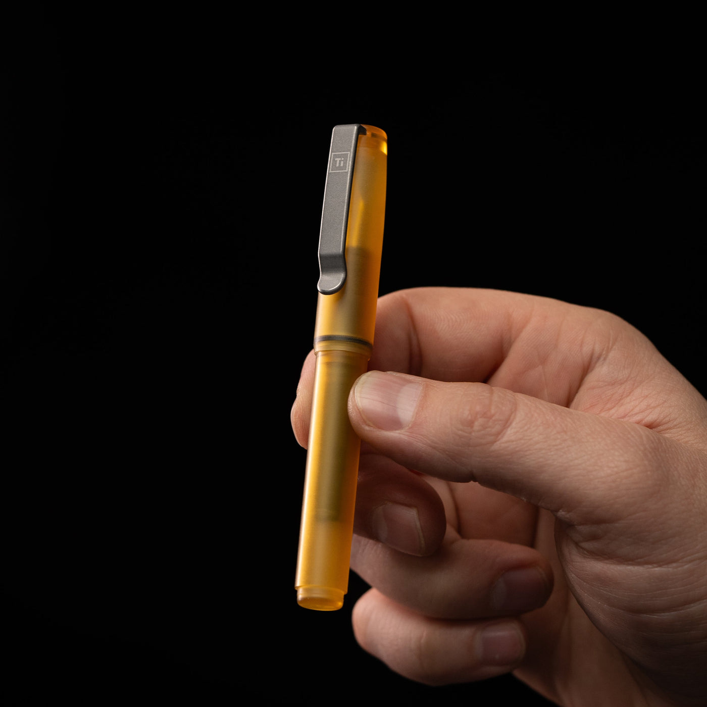 Hand holding a translucent amber USA made EDC pen by BIGiDESIGN-USA with metal clip on black background