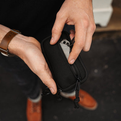 Hands opening a black EDC pocket pouch with a small folding knife inside, outdoors view