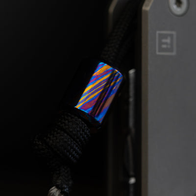 Damascus lanyard bead with vibrant blue and orange patterns attached to black paracord by Big Idea Design LLC