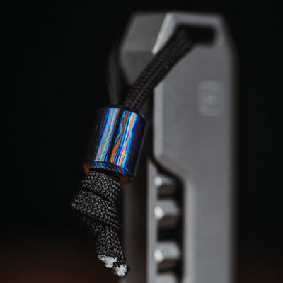Big Idea Design LLC Damascus lanyard bead in blue and orange tones attached to black cord on a metal tool