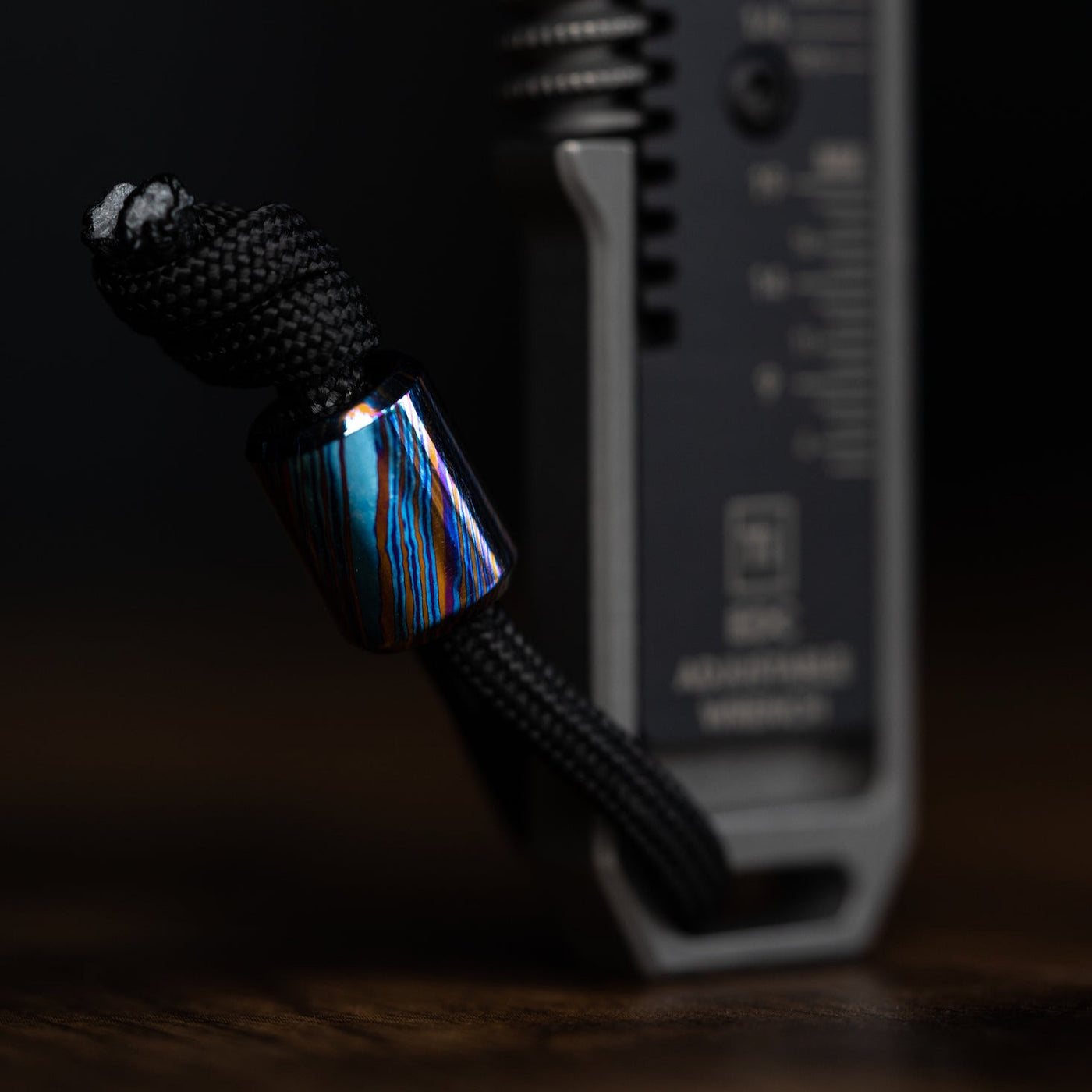 Damascus lanyard bead with blue and brown pattern attached to black paracord on a tool by Big Idea Design LLC