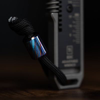 Damascus lanyard bead with blue and brown pattern attached to black paracord on a tool by Big Idea Design LLC