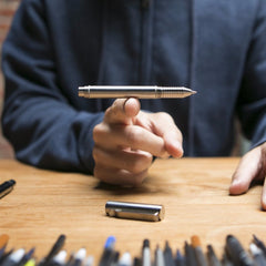 Silver metal EDC refillable pen by BIG IDEA DESIGN LLC being balanced on one finger over wooden table