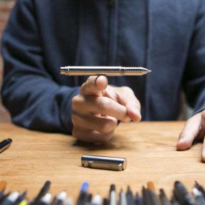 Silver metal EDC refillable pen by BIG IDEA DESIGN LLC being balanced on one finger over wooden table