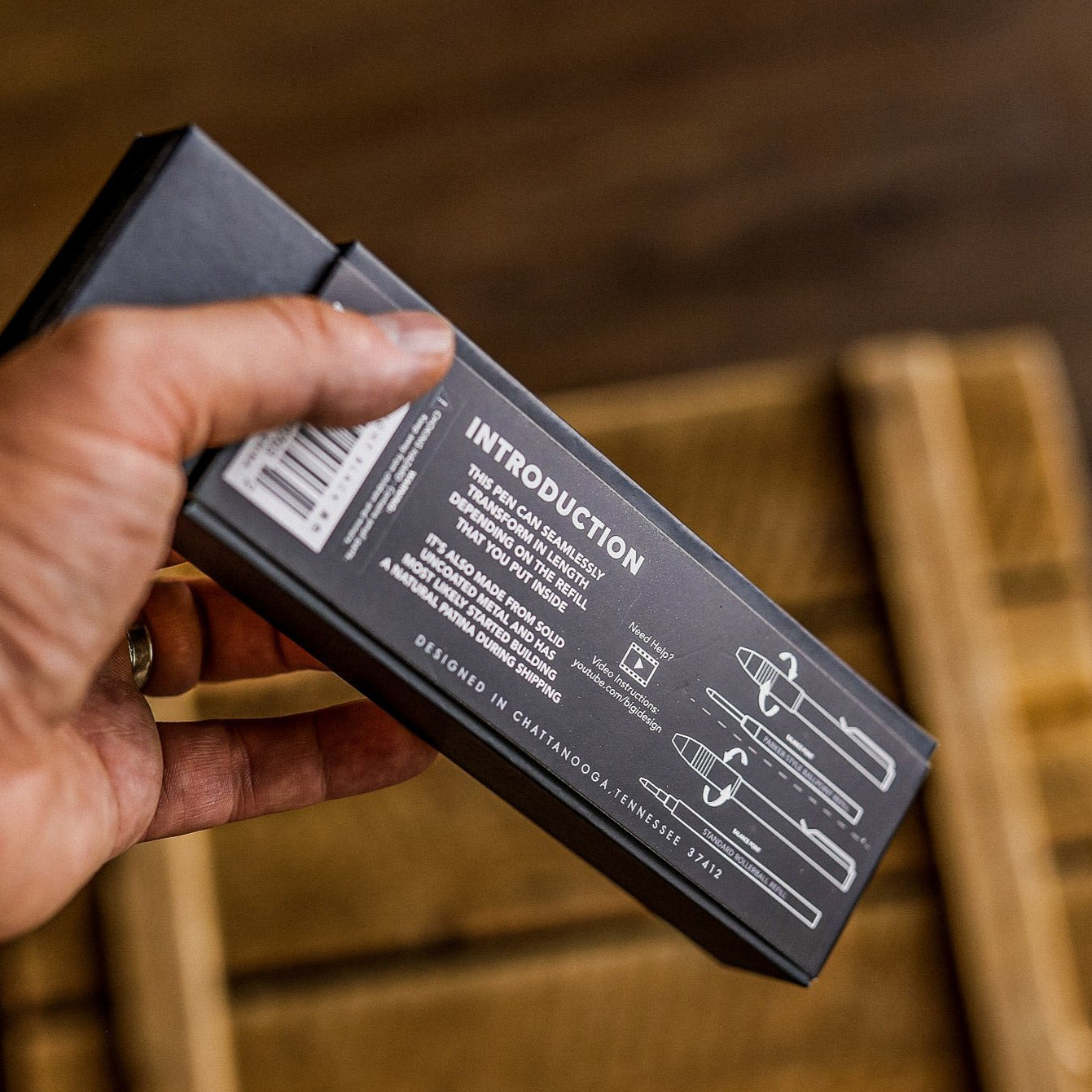 Hand holding BIG IDEA DESIGN pens black box packaging with introduction text and pen diagrams on wooden background