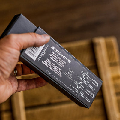 Hand holding BIG IDEA DESIGN pens black box packaging with introduction text and pen diagrams on wooden background