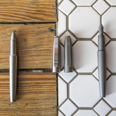 Big Idea Design LLC Titanium Fountain Pen with sleek silver body and two interchangeable caps on rustic wood and white tile background
