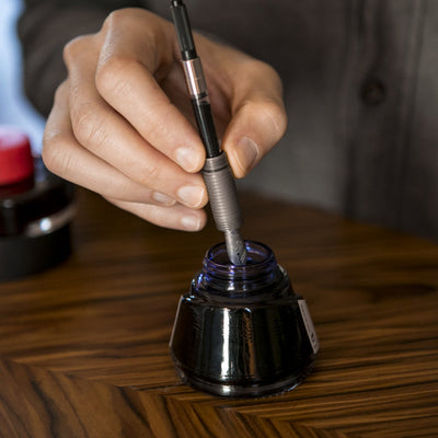 Hand dipping a Titanium Fountain Pen into an ink bottle on a wooden table by Big Idea Design LLC