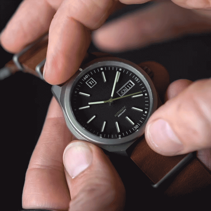 Big Idea Design LLC titanium automatic field watch with black dial and brown leather strap held in hand