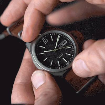 Big Idea Design LLC titanium automatic field watch with black dial and brown leather strap held in hand