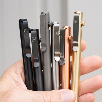 Hand holding six titanium bolt action pens in black, gray, silver, copper, and gold finishes by Big Idea Design LLC