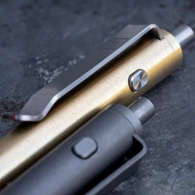 Close-up of two titanium EDC pens by Big Idea Design LLC with metal clips and push buttons on a dark surface