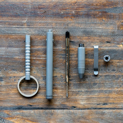 Disassembled titanium EDC pen by Big Idea Design LLC showing all parts on wooden surface