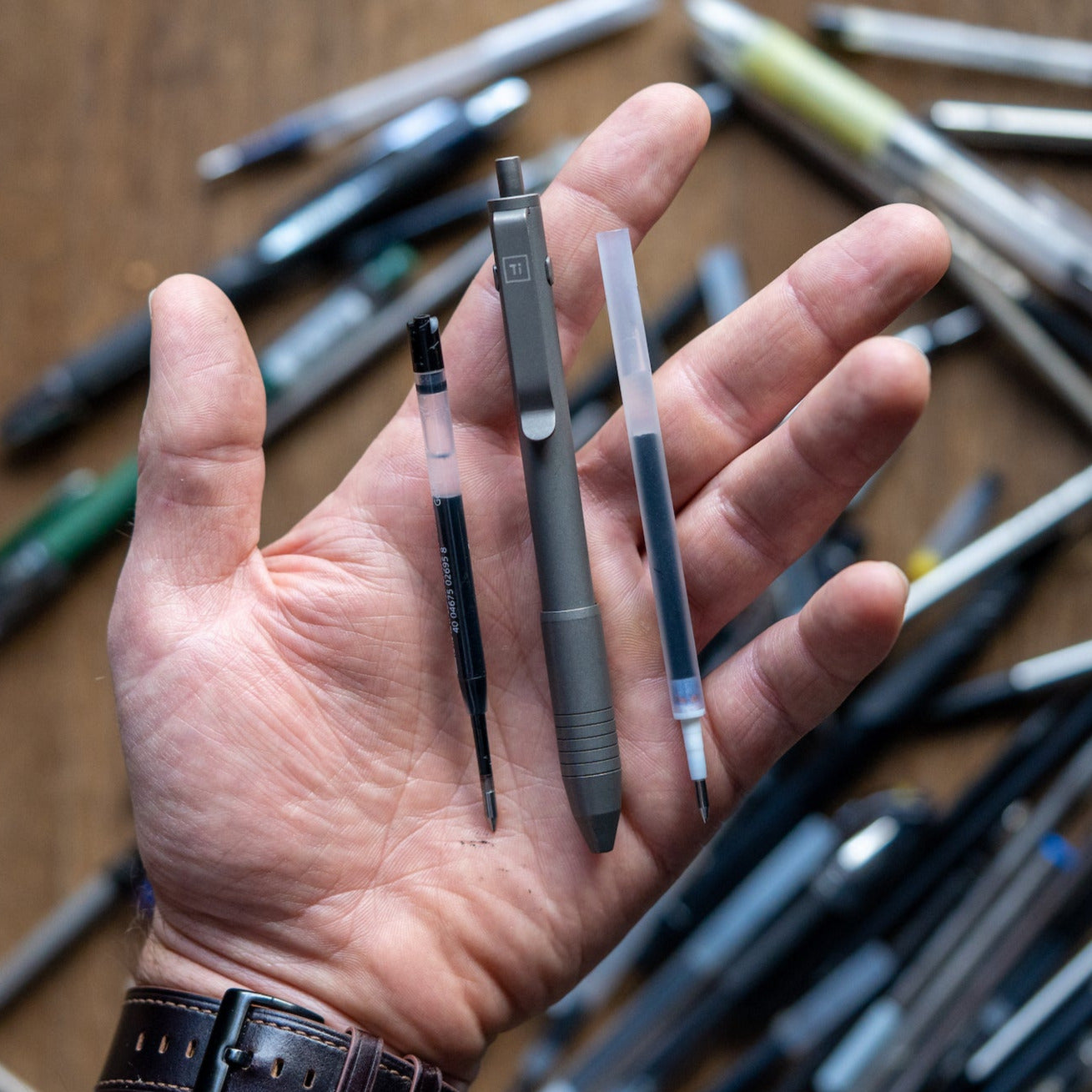 Big Idea Design LLC titanium EDC pen with refill cartridges held in hand over assorted pens