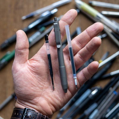 Big Idea Design LLC titanium EDC pen with refill cartridges held in hand over assorted pens