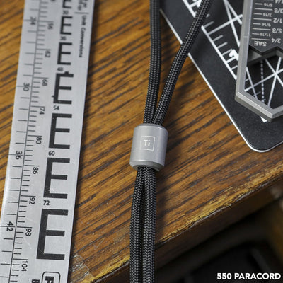 BIG IDEA DESIGN LLC titanium lanyard bead on black 550 paracord next to a ruler on wooden surface
