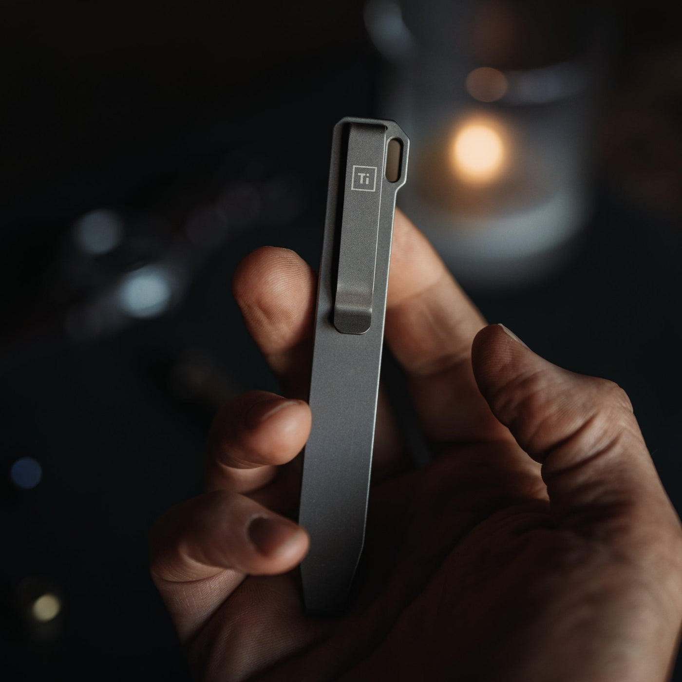 Hand holding a sleek titanium pry bar tool with clip and Ti marking in dim light