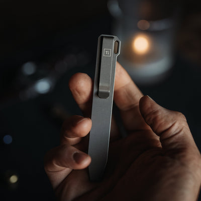 Hand holding a sleek titanium pry bar tool with clip and Ti marking in dim light
